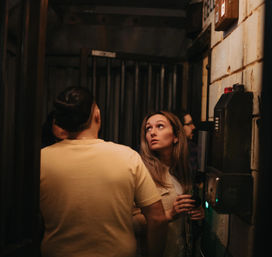 Group of young adults in a dim industrial basement corridor, woman holding a key fob at a wall-mounted keypad while looking upward.