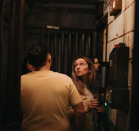Group of young adults in a dim industrial basement corridor, woman holding a key fob at a wall-mounted keypad while looking upward.