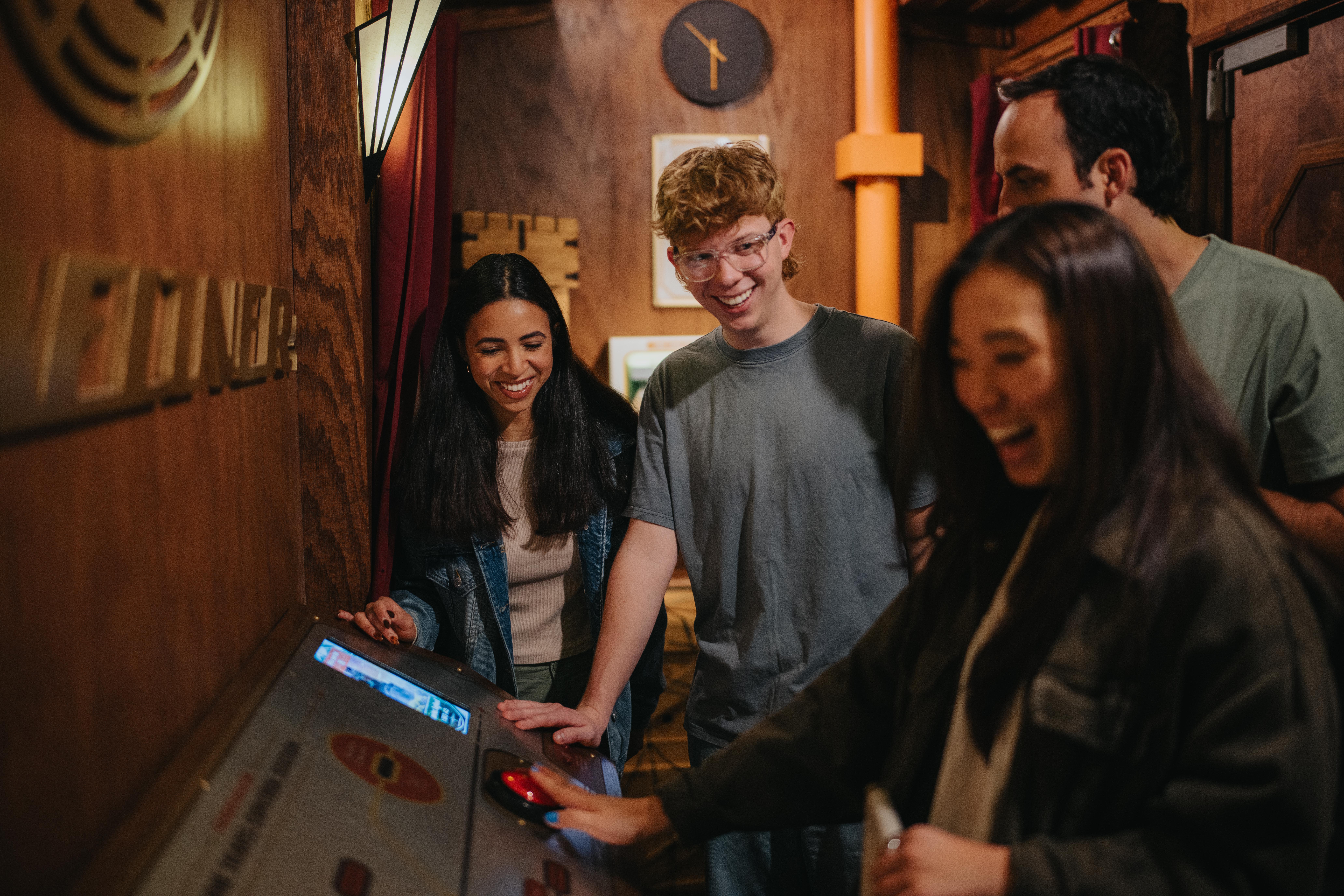 Group of four laughing as they play a retro arcade touchscreen in a cozy wood‑paneled game room with warm lighting