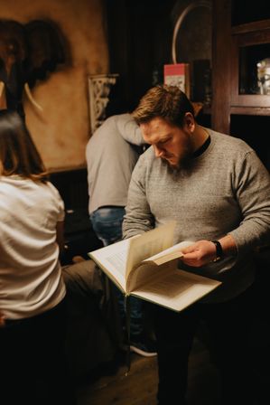 Bearded man flipping through a large hardcover in a cozy, dimly lit bookstore with wooden shelves and elephant wall decor, others browsing in the background.