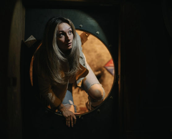 Woman climbing through a round porthole-style doorway in a dim, industrial-lit room, wearing ripped jeans and a jacket with a warm backlight and curious expression.