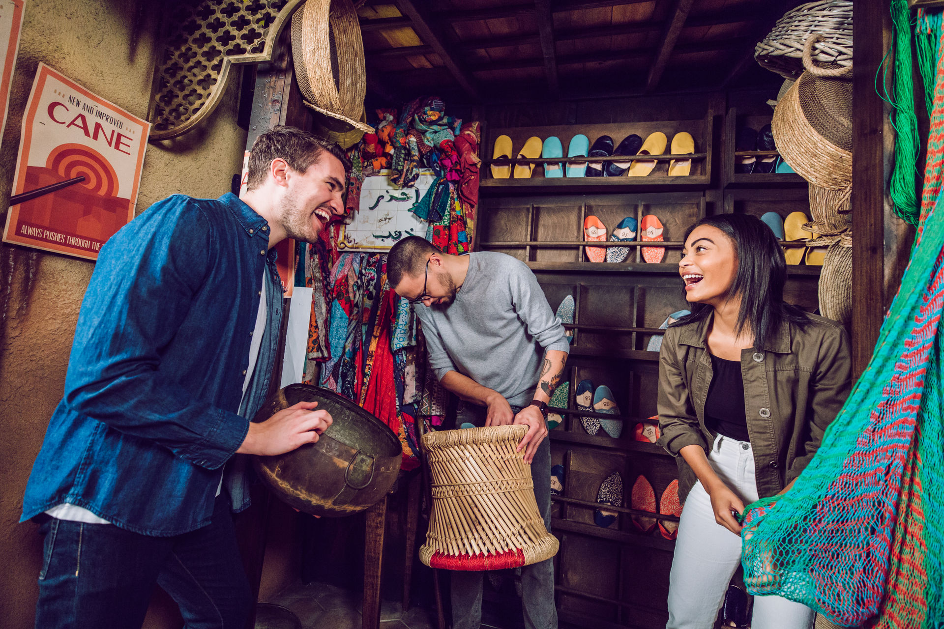 Three young adults laughing and browsing a Moroccan-style market stall filled with colorful slippers, hanging scarves, woven baskets and a handwoven stool.