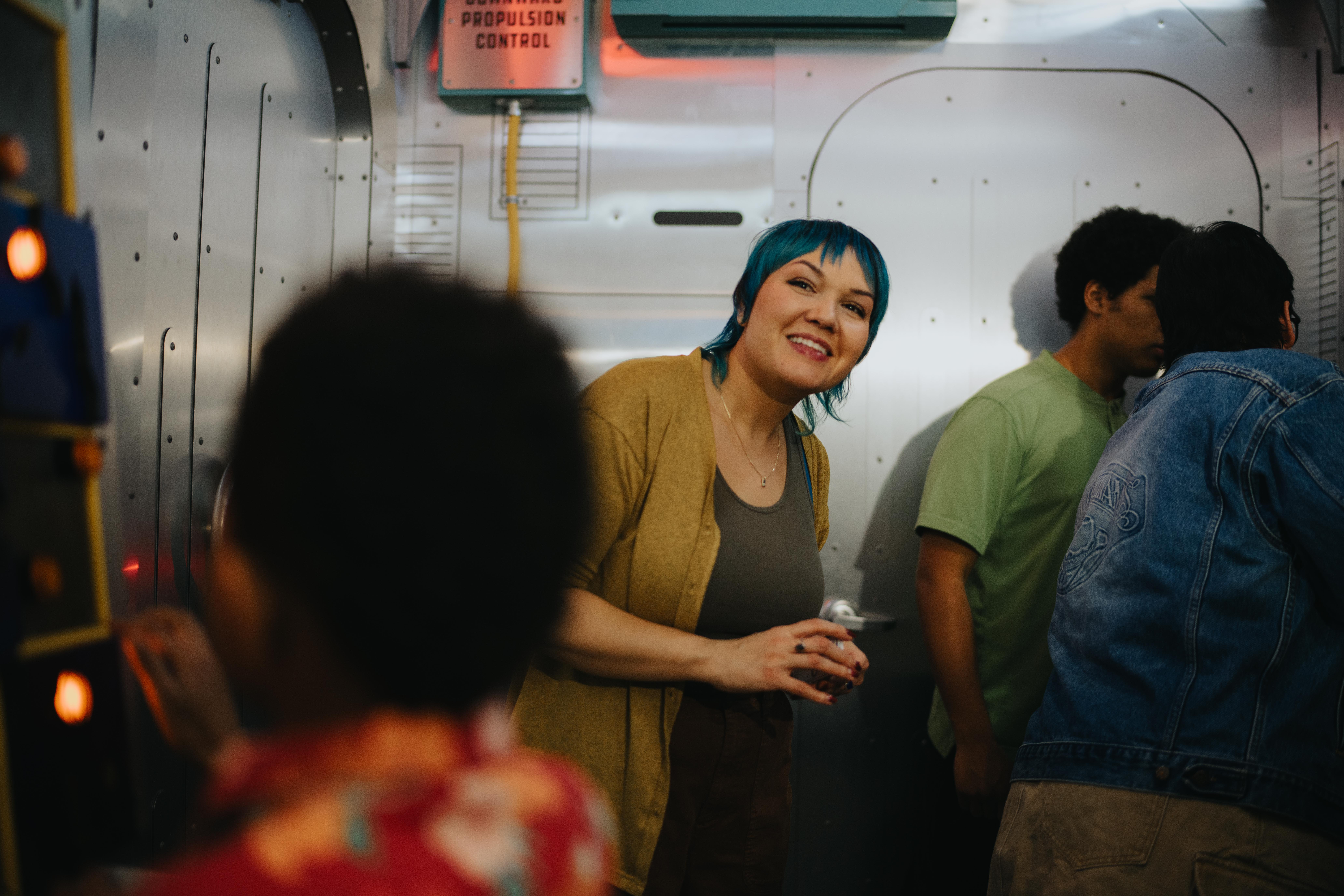 Smiling woman with blue hair leans into a metallic, spaceship-themed interactive arcade exhibit, holding a controller while others use illuminated controls.