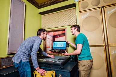 Two men smiling and collaborating in a colorful recording studio control room at a mixing console, one pointing to a computer screen showing audio tracks, surrounded by acoustic panels and large speaker cabinets.
