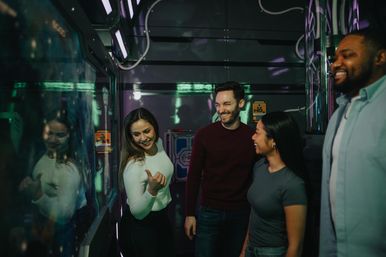 Four friends laughing in a neon-lit indoor game room, woman in a white sweater gesturing at a glowing glass panel while others smile.