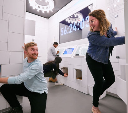 Energetic group of people solving puzzles and opening cabinets in a bright modern escape room with a large countdown timer on the wall.