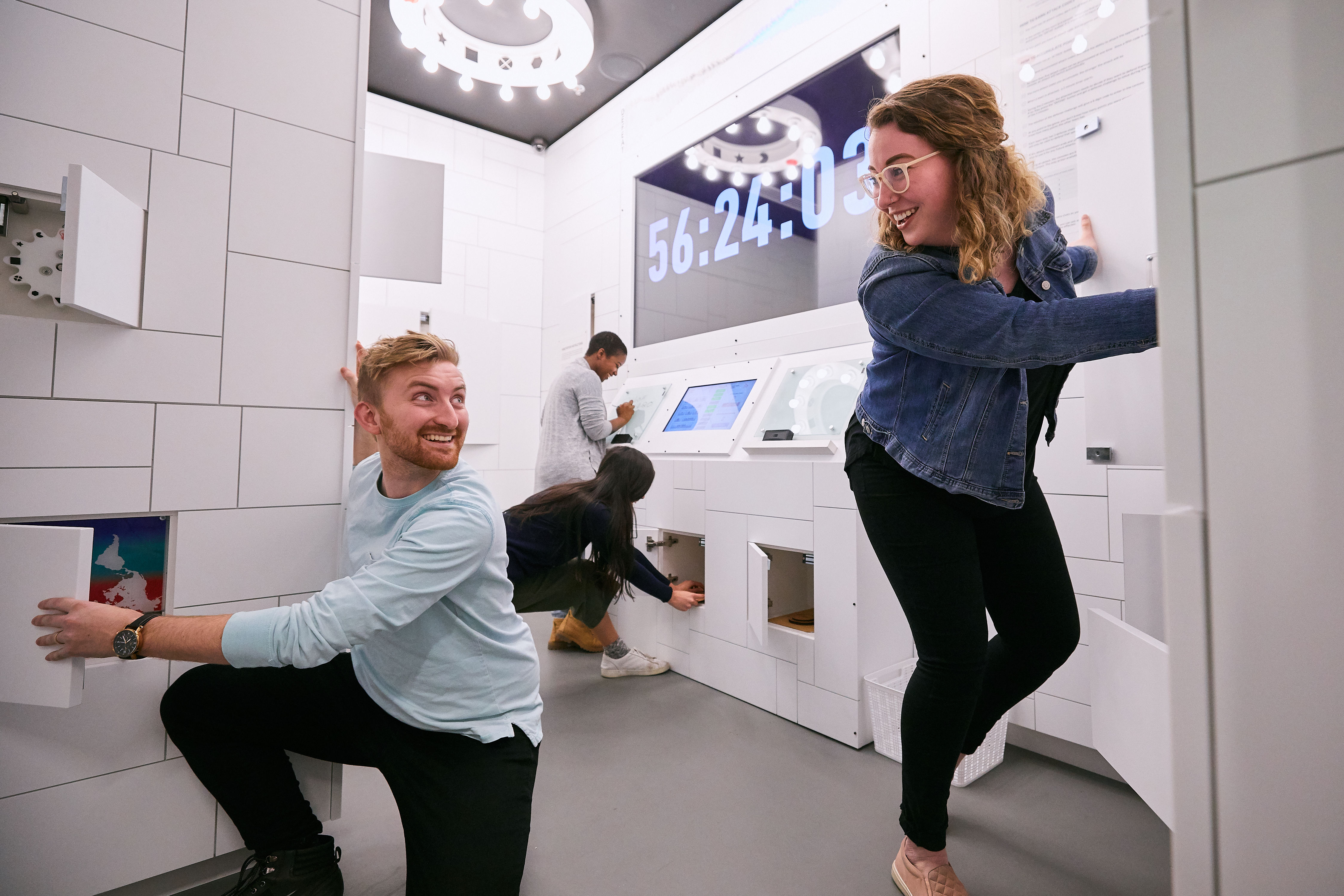 Energetic group of people solving puzzles and opening cabinets in a bright modern escape room with a large countdown timer on the wall.