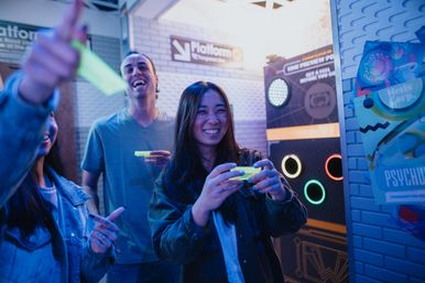 Smiling friends play a neon-lit interactive arcade game in an indoor amusement area, holding glowing controllers and aiming at colorful lighted targets.