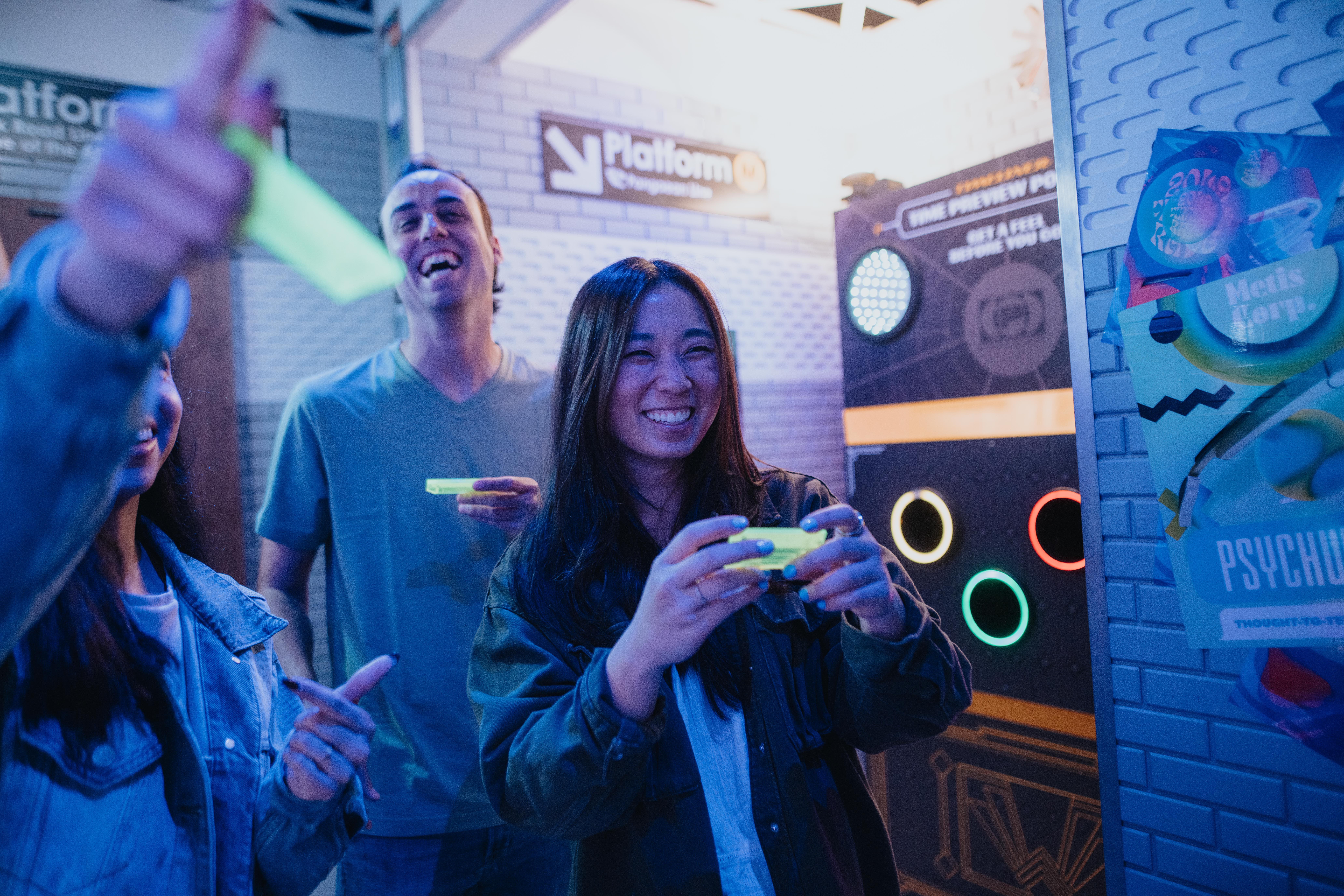 Group of young adults at an indoor arcade playing a neon-lit target game — smiling woman holding a glowing controller with colorful circular targets and posters in the background