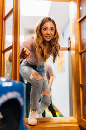 Young woman in ripped jeans and white sneakers crouches through a wooden-framed glass door into a bright indoor hallway, smiling and reaching forward.