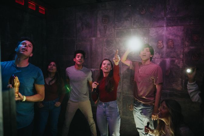 Excited group of young adults in a dim stone-walled escape room, using flashlights and pointing at a clue during a puzzle-solving adventure.