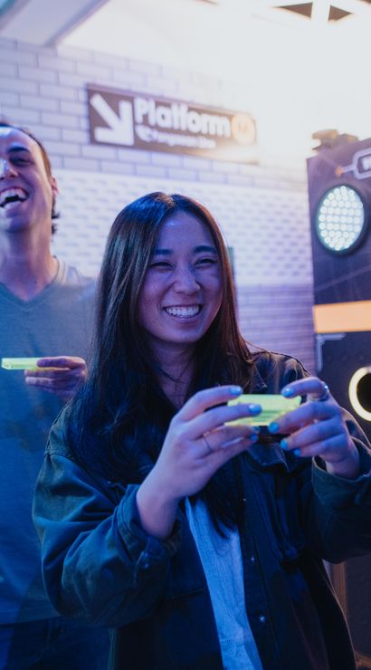 Young friends playing a neon-lit arcade game indoors — smiling woman holding a glowing controller while colorful circular targets light up on the game cabinet.