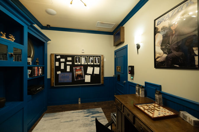 Cozy blue-trimmed interior with built-in shelving, a bulletin board pinned with photos and notes, a wooden desk with a Scrabble-style tile board, rug on dark hardwood floor, and a framed poster on the wall.