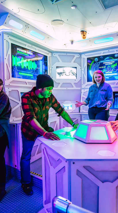 Group of four in a space-themed escape room gathered around a glowing control console and futuristic wall screens under purple and blue neon lighting, solving an interactive sci‑fi puzzle.
