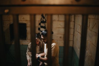 Two people behind metal bars in a dim, cell-like room, inspecting handcuffs by worn concrete walls and a hanging striped garment — moody, cinematic escape-room or vintage jail vibe.