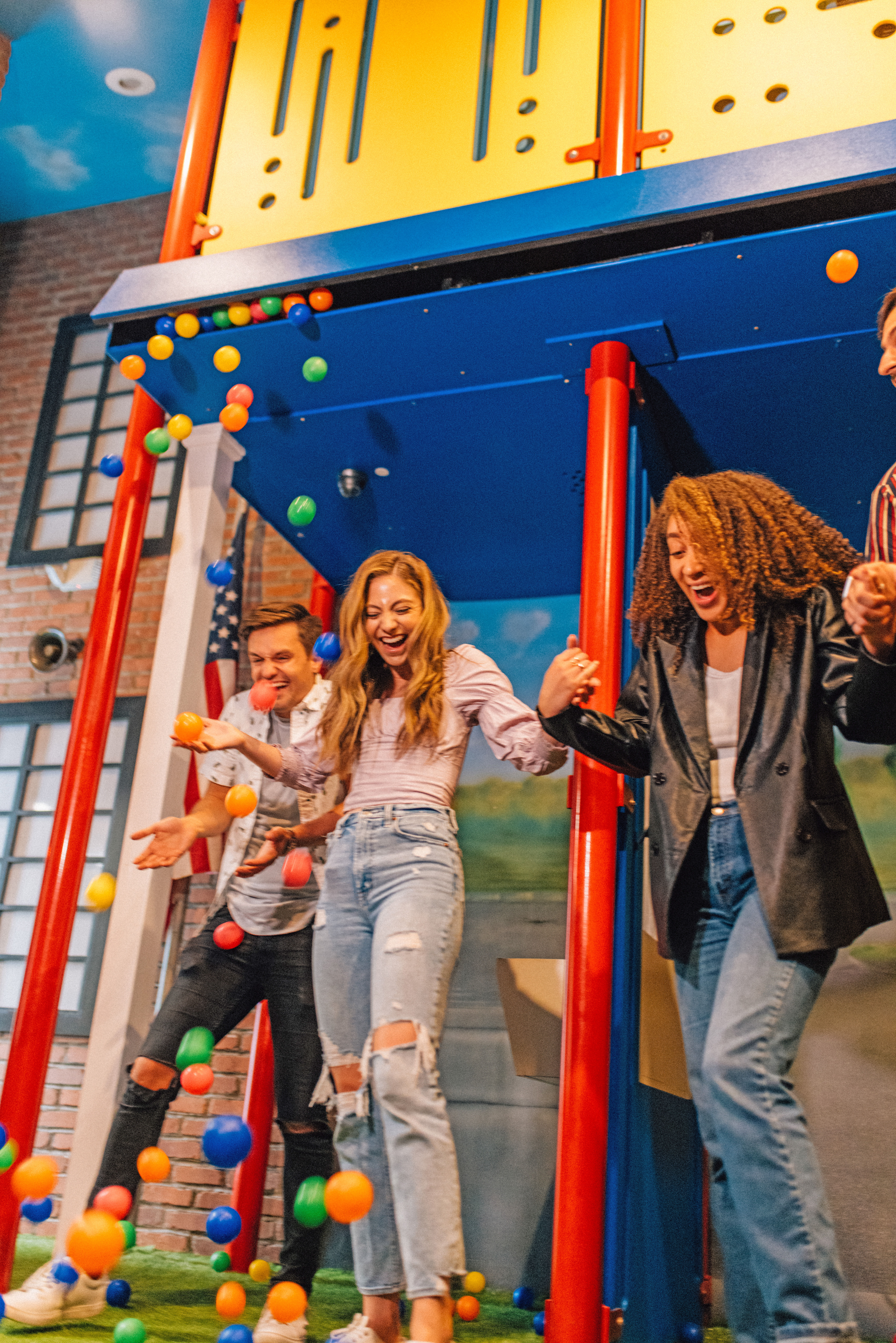 Three friends laughing and holding hands beneath a bright blue-and-yellow indoor play structure as colorful plastic balls cascade down in a lively indoor playground.
