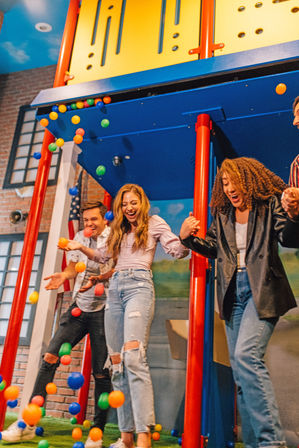 Three friends laughing and holding hands beneath a bright blue-and-yellow indoor play structure as colorful plastic balls cascade down in a lively indoor playground.
