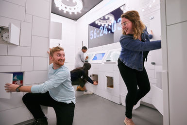 Energetic group of people solving puzzles and opening cabinets in a bright modern escape room with a large countdown timer on the wall.