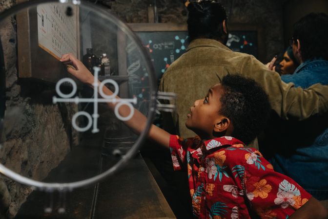 Curious child in a red floral shirt reaches to touch an interactive science exhibit through a circular glass panel in a dim, stone‑walled museum gallery with adults observing in the background.