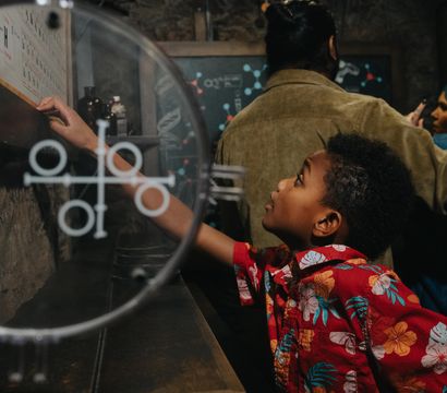 Curious child in a red floral shirt reaches to touch an interactive science exhibit through a circular glass panel in a dim, stone‑walled museum gallery with adults observing in the background.