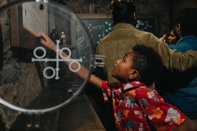 Curious child in a red floral shirt reaches to touch an interactive science exhibit through a circular glass panel in a dim, stone‑walled museum gallery with adults observing in the background.