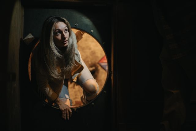 Young woman climbing through a round porthole-style doorway into a dim, warmly lit indoor room, wearing ripped jeans and a jacket and looking up with a curious expression.