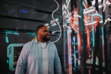 Smiling man in a light-blue shirt exploring a neon-lit, futuristic tech exhibit with glowing glass tubes and colorful reflections