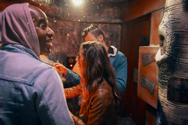 Three friends crowded in a dimly lit escape room, examining clues on a rustic wall under a single hanging bulb — lively indoor puzzle-solving scene.
