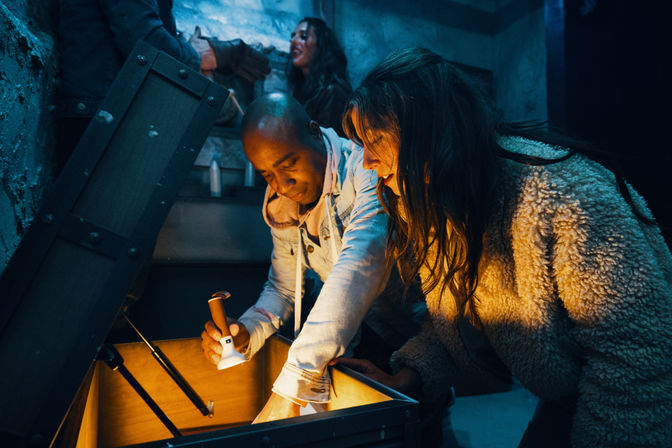 Two people leaning over an open wooden chest in a dim, industrial-style room, one holding a flashlight that lights the chest’s interior while another figure watches in the background — suspenseful treasure-hunt/escape-room vibe.