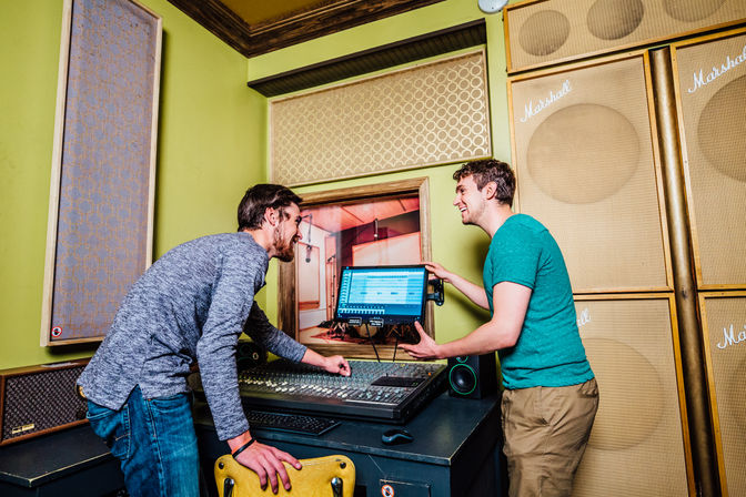 Two men smiling and collaborating in a colorful recording studio control room at a mixing console, one pointing to a computer screen showing audio tracks, surrounded by acoustic panels and large speaker cabinets.