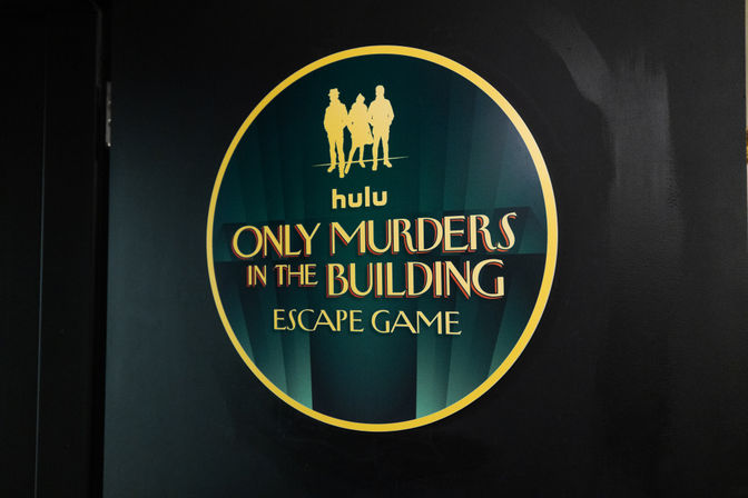 Round art-deco wall sign for a murder-mystery escape game, emerald-green and gold design with three silhouetted figures.
