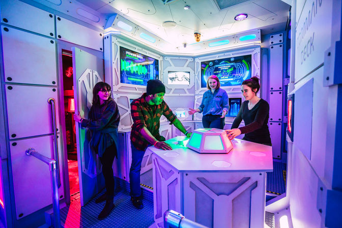 Group of four in a space-themed escape room gathered around a glowing control console and futuristic wall screens under purple and blue neon lighting, solving an interactive sci‑fi puzzle.