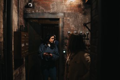 Two people explore a dim brick-walled industrial utility room with pipes, pressure gauges, a control panel and a security camera under moody warm lighting.