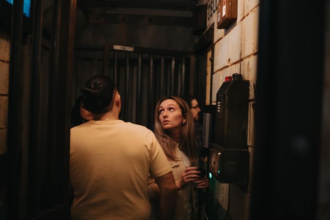 Young adults in a narrow, dim industrial-style corridor with metal bars and a wall-mounted payphone/intercom; a woman holds the receiver and looks up, scanning the space.