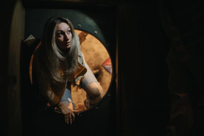 Woman in ripped jeans climbing through a round metal hatch into a dimly lit, warmly backlit room, looking up with a curious gaze