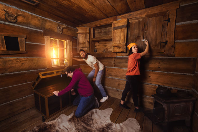 Three people in a rustic log cabin escape room: one kneeling to open a wooden chest, one unlocking a wall cabinet, and one peering toward a sunlit window; wood stove, mounted antlers, and cowhide rug in the room.