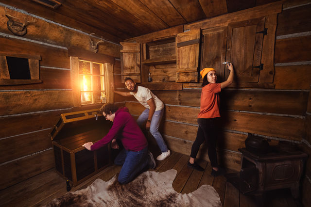 Three people in a rustic log cabin escape room: one kneeling to open a wooden chest, one unlocking a wall cabinet, and one peering toward a sunlit window; wood stove, mounted antlers, and cowhide rug in the room.