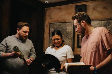 Three adults in a cozy dim room examining clues: one holding a wine bottle, another reading an open book, and a smiling person inspecting a black hat in front of framed vintage maps.