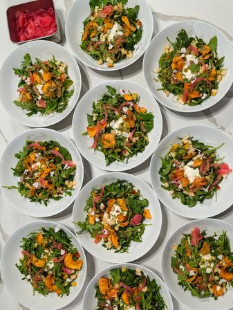 Top-down view of multiple white salad bowls on a marble countertop: fresh arugula with creamy burrata, roasted sweet potato chips, pickled red onions, diced apple, edible flowers and herb vinaigrette.