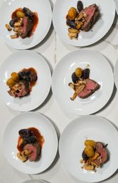 Top-down view of multiple white plates with medium-rare beef, roasted yellow and purple baby potatoes, mixed mushrooms and glossy red‑wine jus on a marble countertop — restaurant-style plated entrees.