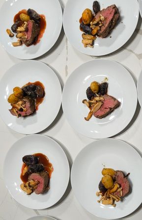 Top-down view of multiple white plates with medium-rare beef, roasted yellow and purple baby potatoes, mixed mushrooms and glossy red‑wine jus on a marble countertop — restaurant-style plated entrees.