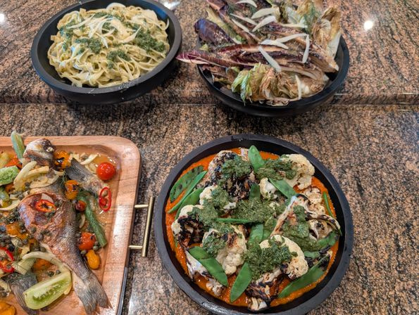 Top-down shot of a restaurant spread on a granite counter: black bowls with pesto fettuccine and charred radicchio salad, a skillet of grilled cauliflower and sugar snap peas on red pepper puree with herb pesto, and a wooden board with whole roasted fish, cherry tomatoes and capers.
