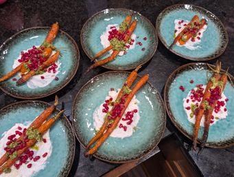 Six turquoise plates of Mediterranean-style roasted baby carrots topped with green herb pesto, creamy labneh and bright pomegranate seeds on a dark stone countertop