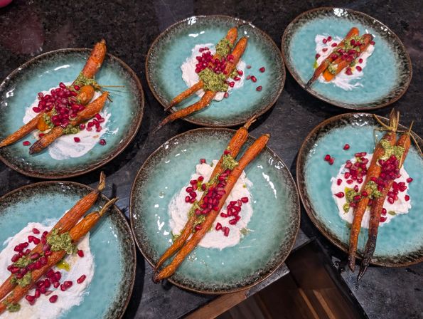 Six turquoise plates of Mediterranean-style roasted baby carrots topped with green herb pesto, creamy labneh and bright pomegranate seeds on a dark stone countertop
