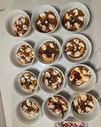 Top-down view of multiple individual creamy dessert cups in white bowls on a marble countertop, topped with cocoa powder, chocolate shavings, tiny meringue kisses and edible flower petals — plated and ready to serve.