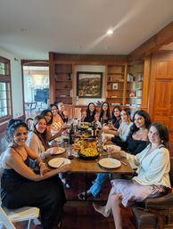 Group of women smiling around a long wooden dining table in a warm home dining room, sharing wine and platters of roasted vegetables and potatoes, with built‑in wood shelves and white balloons visible in the adjacent room.