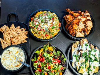 Top-down view of a vibrant Mediterranean-style spread on a dark table: grilled chicken, pearl couscous salad with feta and herbs, mixed summer salad, roasted eggplant and zucchini with yogurt drizzle, creamy tzatziki dip and toasted flatbread slices in black bowls.