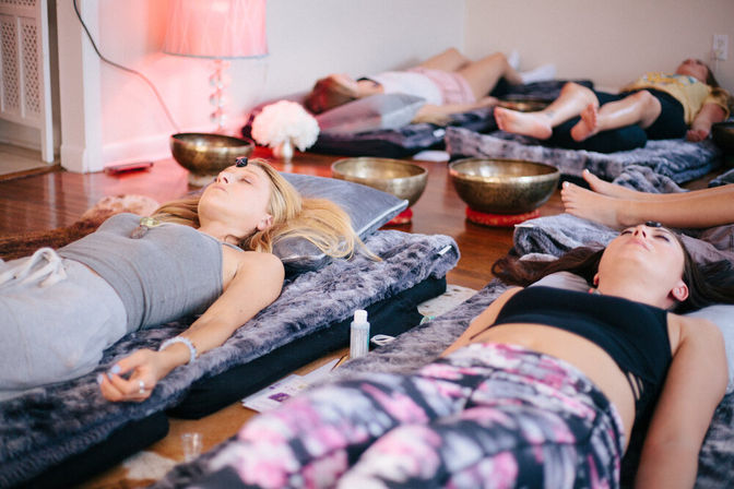 Group sound bath in a cozy wellness studio — people reclining on cushioned mats with singing bowls and forehead crystals for guided meditation.