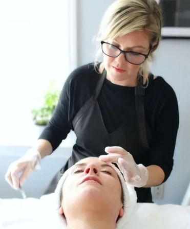 Professional esthetician in glasses and gloves applying a soothing facial treatment to a relaxed client on a bright spa bed in a skincare treatment room.