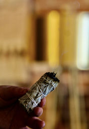 Hand holding a tied white sage smudge stick with a charred tip against a warm, blurred indoor background — smudging/cleansing ritual